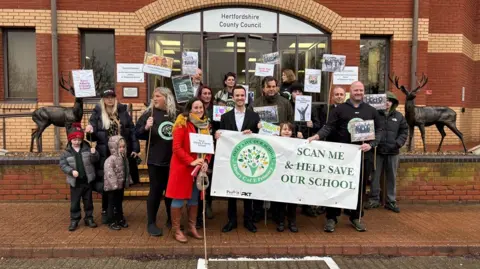 A group of adults and children posing outside the brick-built offices of Hertfordshire County Council in Stevenage. They are holding signs and a large white banner.