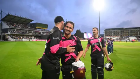 Getty Images Lewis Gregory and Craig Overton of Somerset celebrate together on the pitch following the Vitality Blast Men match between Somerset and Middlesex in Taunton. They are in the county's black and pink kit and in the background the full stands and stadium floodlights are visible