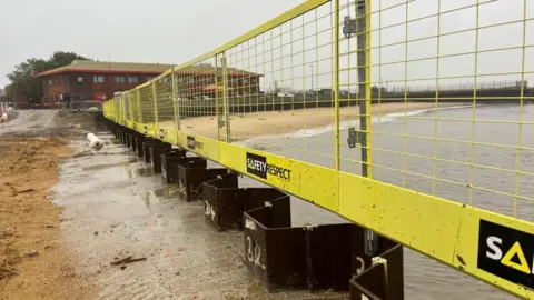 Andrew Turner/BBC Sheet steel piling and concrete with yellow fencing and a beach on the river in the distance