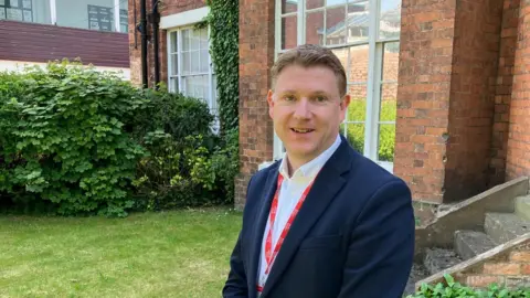 A man with short brown hair, a blue suit and red tie stood in front of a brick building
