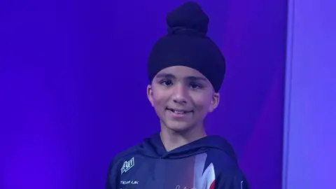 A young boy smiling at the camera. he is wearing a turban and a sports hooded top and in a room with purple walls.
