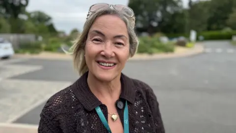 A woman with fair hair held back by glasses on her head is smiling at the camera. She is standing against the backdrop of a car park, and is wearing a brown cardigan and blue hospice lanyard.