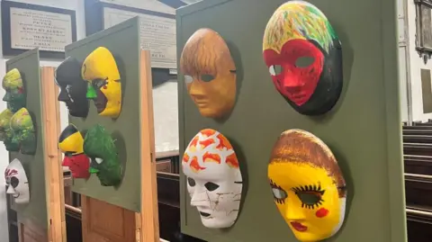Maddy Jennings/BBC 11 masks, handing from a green board. Each mask is different, painted with vibrant colours and patterns.