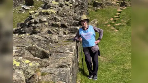 Family handout A woman in hiking gear, a green brimmed hat and a blue vest leans on a section of Hadrian's Wall. She is carrying a hiking pole.