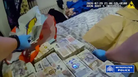 Bodycam footage showing gloved hands handling large bundles of cash on a bed, with Metropolitan Police watermark and timestamp.