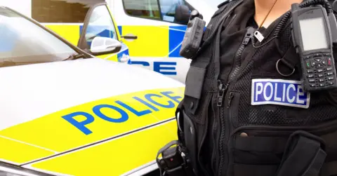 Getty Images The front of a police car and an office standing next to it. On the officer's chest is a blue strip saying "police" in white behind a walkie-talkie. There is another police car behind the first
