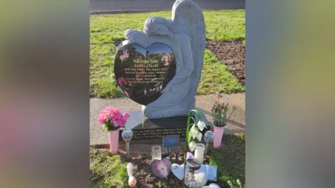 Family A grave marked with a carved stone angel holding a heart. Flowers and pictures surround it