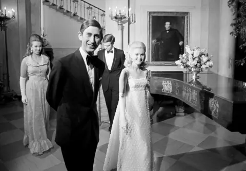 AP Princess Anne, Prince Charles, David Eisenhower and Tricia Nixon smile as they look at the camera while walking through a room in the White House in formal attire, in a black and white photo.