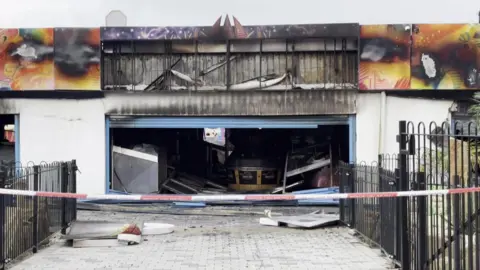The arcade at Ocean Beach Pleasure Park after the fire. The metal shutters are open and show damage inside the building. Sheets of metal have fallen to the floor. Red and white no entry tape has been hung across the entrance.