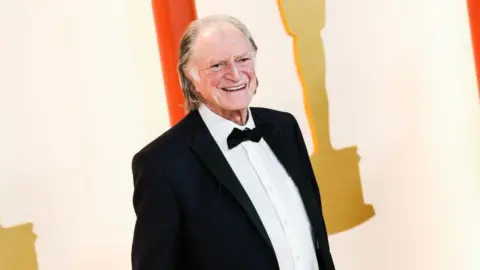 Getty Images An elderly man with white hair brushed back poses in black tie on a red carpet in front of a white backdrop with images of golden statuettes on it.