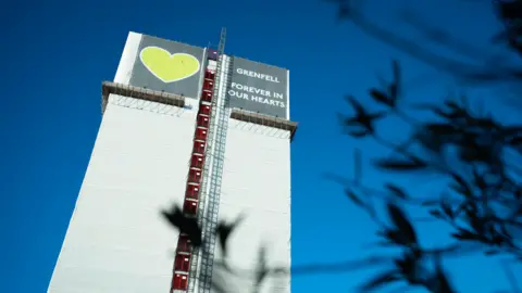 PA Media Grenfell Tower wrapped in white sheeting, with a large banner at the top displaying a green heart and the words "Grenfell forever in our hearts." The sky is clear and blue, and blurred dark leaves frame the right side of the image.