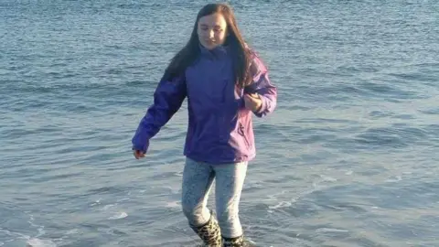 Williamson Family Rachel is walking on shallow sea water in black and white wellie boots. She is wearing denim jeans and a purple short coat. Her hair is long and brown. 