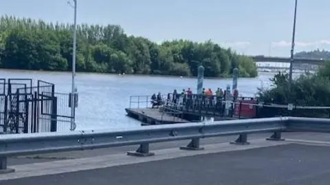 A large body of water with a platform on which several people are stood in a crowd. In the background are trees and in the foreground is a road with a metal barrier.