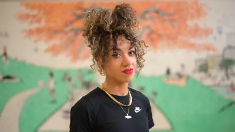 Alexander Polis Jasmine has blonde and brown curls tied on top of her head. She is wearing a black Nike t-shirt and is smiling at the camera.