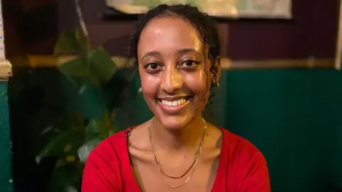 Selam Haile is wearing a red top and two gold necklaces. She has black curly hair and is smiling at the camera. She is sitting in front of a tall green plant and a brown and green wall.