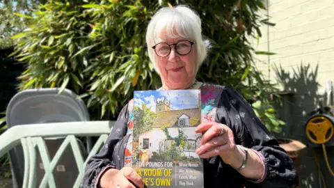 Sarah Hosking sitting in her garden and holding her new book 'Five Pounds for a Room of One's Own'.