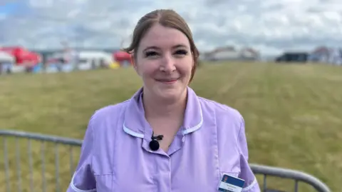 A woman with reddish-brown hair pulled back and long eyelashes in a light purple nursing style top with a name badge stands in front of a fence with a showfield behind her