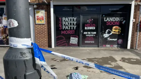 BBC The front of the takeaway can be seen across the wide pavement outside. There are promotional posters stuck to the windows that tell people the burgers it will be producing are "landing soon". They also say "Hello Bloxwich". In the foreground police tape can be seen tied to a lamp-post.