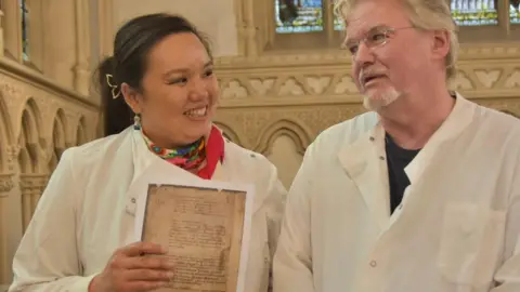 Corpus Christi College, Cambridge Dr Betty Chung with dark hair pulled back in a pony tail and a red neck scarf, holding a copy of the 9th Century manuscript page, and Prof John Carr with white hair and beard and wearing rectangle glasses, both wearing white lab coats in Corpus Christi College chapel