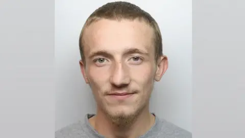 A mugshot of a man with a short brown hair, beard and moustache. He is wearing a grey top and is looking straight into the camera.