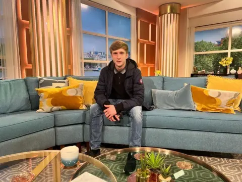Matthew sits on This Morning couch. He wears jeans, a long sleeve dark polo top and a dark jacket. He has blonde hair which is styled in a quiff.