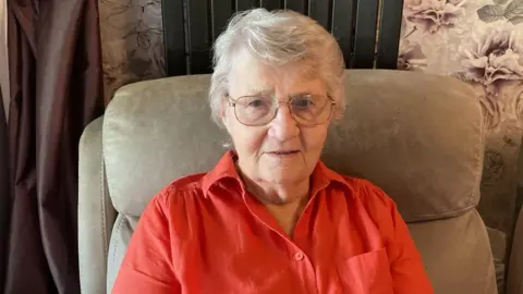 Picture of older woman sitting in chair and facing the camera. She is wearing a red shirt and glasses. 