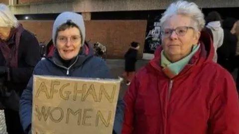 Two women, one in a blue coat and a hat holding a sign that reads 'Afghan women'. The other woman is in a red coat, and she has short white hair