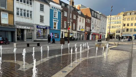 Alex Pope/BBC Northampton Market Square is seen from ground level. Jets of water are coming from a paved surface as part of a water feature. Rows of shops, including a Coral betting shop and a bakery, can be seen.