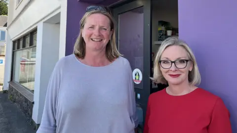 Jennifer Merrett, left, is a woman wearing a grey bowl-neck jumper. She is smiling and has blue eyes and fair hair. She has sunglasses on her head and is stood outside the purple walls of the salon. Racheal Day, right, is a petite woman with short bobbed blonde hair, black glasses and red lipstick. She is stood smiling and wearing a red dress.