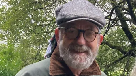 A man with a beard and glasses, wearing a grey flatcap