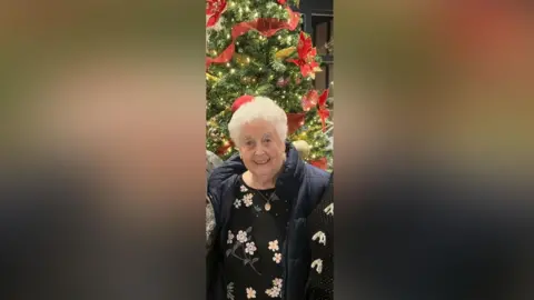 Family Handout A woman in a black top and blue coat. She has grey hair and is standing in front of a Christmas Tree.