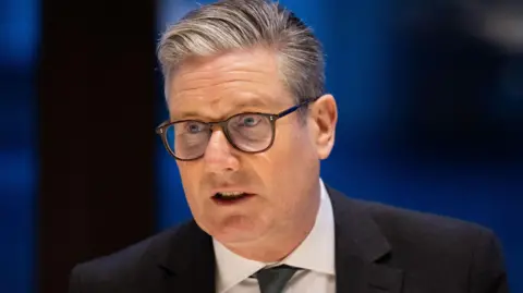 Sir EPA Keir Starmer, wearing jacket and tie, glasses, in front of a dark blue background