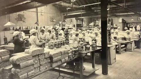 Hull History Centre An old photo, in black and white. It shows a group of about 30 factory workers all wearing white bonnets and white aprons. They are all gathered around different tables which have clear glass jars of sweets on them. There are also lots of parcels at the forefront of the photo.