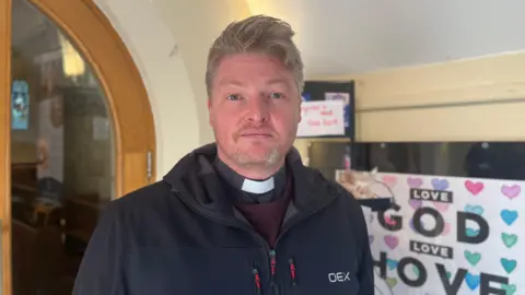 Josh McLaughlin/BBC Revered Dan Henderson, wearing his vicar's collar, faces the camera while standing in an indoor entryway to the St Andrew's Church in Hove. A decorative sign reading "LOVE GOD, LOVE HOVE" hangs on the wall behind him, embellished with multi-colour hearts.