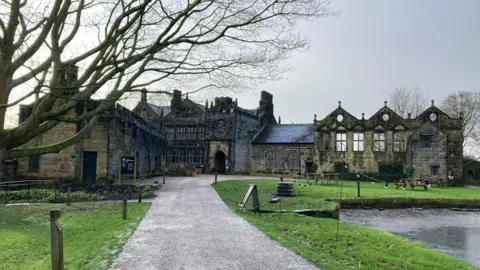 Charles Heslett/BBC A partially ruined stone manor house, set in grassy surroundings with a pond to the right and a tree to the left and a gravel path leading up to its entrance