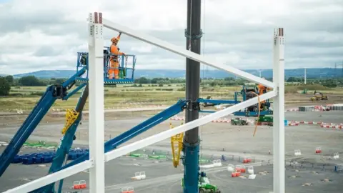 Agratas A photo of a large steel frame being erected on a building site with a construction worker dressed in orange high-vis clothing on a crane next to it.