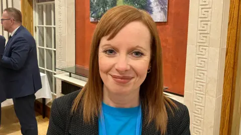 Alma White - a woman with shoulder-length, auburn hair looks at the camera. She is wearing a blue t-shirt and a black and white polka dot suit jacket.