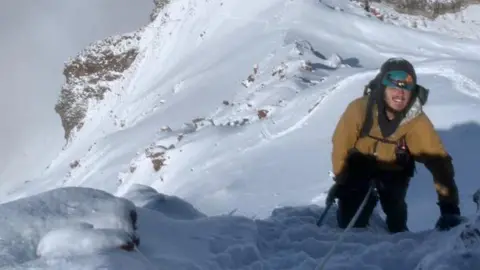 Ollie Treviso A man scaling a snow covered mountain top. He attached to a rope and he's wearing a helmet and large jacket.