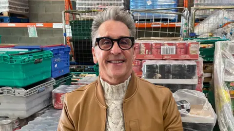 Neil Reid founder and chairman of The Big Food Project with grey hair wearing black rimmed glasses and a cream jumper and tan-coloured leather jacket in a warehouse of food