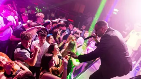 Shad Ellis Shad is on stage dressed as Men In Black Will Smith and performing for a smiling crowd of young people with a microphone in his hand.