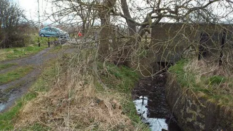 The tiny stream at Cut Throat Dene in Sunderland. The water is murky with leaves floating in it. A tree is above the stream, which is next to a path. A blue car is parked at the top of the stream.
