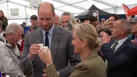 Prince William and Duchess of Edinburgh chink paper cups together as they sample an alcoholic beverage at the Royal Cornwall Show. They are inside a large white marquee. A large crowd of people has surrounded them.