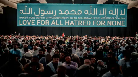 A crowd of people sat in a building under a large banner which says "There is none worthy of worship except Allah Muhammad is the messenger of Allah" "Love for all Hatred for none"