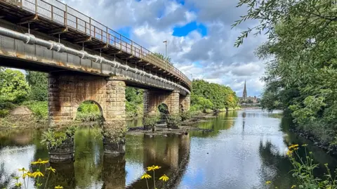 Brian Johnston A bridge over water, stretching into the distance. A church and its spire tower can be seen in the background. 