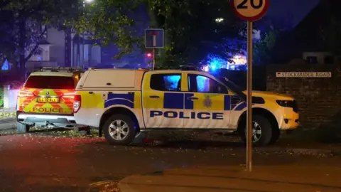 Two police vehicles parked on a road. There are blue lights in the background.