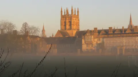 Lucie Johnson An Oxford college appears through the mist in the morning sun. The tower and spires are glowing in the early morning light. In the foreground a playfield is covered in a light mist