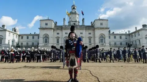 PA Pipe Major Scott Methven in Highland dress including kilt, playing the bagpipes, in front of assembled rows of military.