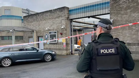 A police officer stands with his back to the camera and is stood near a grey, unmarked police car. There is a police cordon closing off the station and a white Audi car in the distance in the station.