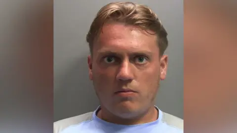 A mugshot of Bradley Cowan. He has short blond hair and blue eyes. He is looking straight at the camera with a serious expression. He is wearing a blue and beige t-shirt.