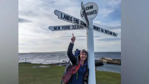 Rebecca Vials A woman wearing a headband, a raincoat and black leggings is standing smiling with her arm up next to a large white sign for John O'Groates. She is on grass and the sea is behind her.